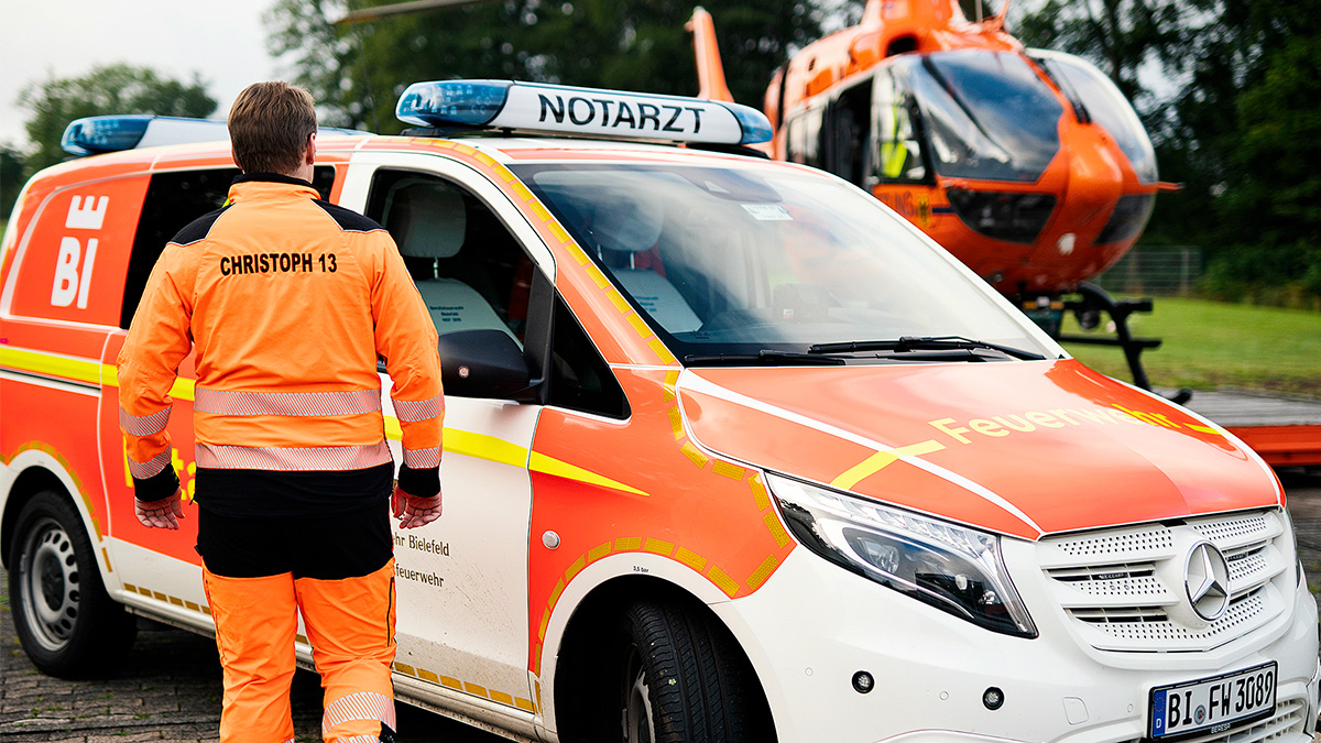 Bild vom Rettungssanitäter vor dem Notarzt Fahrzeug im Hintergrund der Hubschrauber Christoph 13